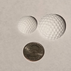 May include: Two white golf balls with dimpled surfaces, one smaller than the other, next to a U.S. quarter coin. The golf balls are on a white surface. The quarter shows the profile of George Washington.
