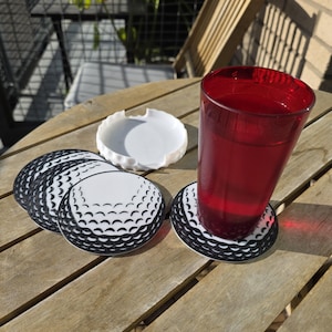 Puede incluir: Un juego de posavasos con temática de pelota de golf con un diseño en blanco y negro, un cenicero blanco y un vaso de líquido rojo sobre una mesa de madera. Los posavasos son redondos y tienen un patrón de hoyuelos.