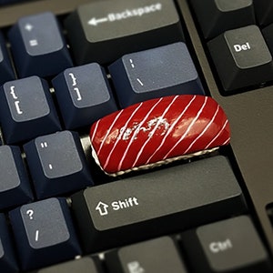 May include: A red and white sushi-shaped keycap sits on a black keyboard with white lettering. The keycap is on the "Shift" key.
