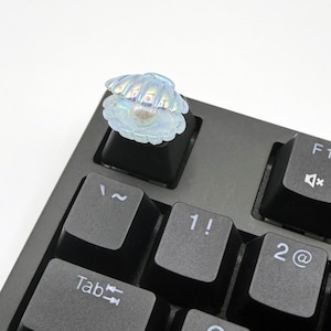 May include: A black mechanical keyboard with a keycap featuring a white seashell with a pearl inside. The keyboard has keys labeled with letters, numbers, and symbols. The seashell keycap is on the "1" key.