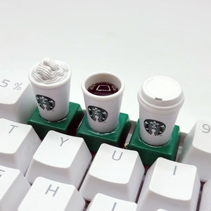Può includere: Tre tazzine da caffè in miniatura su una tastiera bianca. Le tazzine sono verdi e bianche con il logo Starbucks. Una tazzina ha panna montata, una caffè e una caffè freddo.
