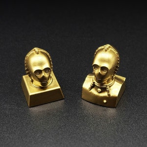 May include: Two gold-colored keycaps featuring the head of C-3PO from Star Wars. The keycaps are designed to fit a standard mechanical keyboard.