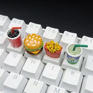 Op de afbeelding: Een wit toetsenbord met vier miniatuurvoedingsmiddelen die op de toetsen zijn geplaatst. De items zijn een hamburger, frietjes, een blikje frisdrank en een glas ijsthee.