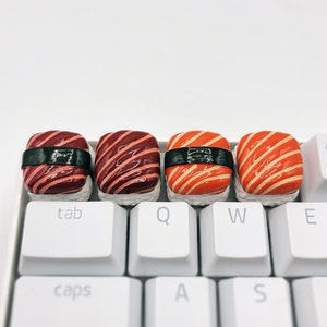 May include: Four miniature sushi keycaps in red and orange with black accents. The keycaps are arranged on a white keyboard with keys labelled 'tab', 'q', 'w', 'e', 'caps', 'a', 's', and 'd'.