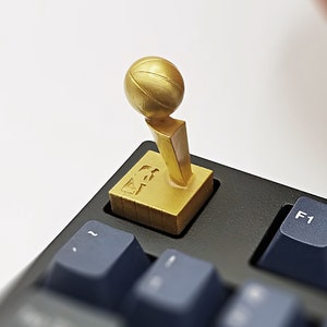 May include: A gold basketball trophy keycap with a basketball on top. The keycap is on a black keyboard with a blue F1 key.