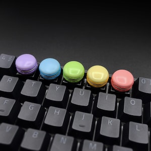 以下が含まれることがあります： 黒いキーボードの上に並んだ5つのカラフルなマカロン。マカロンは、紫、水色、緑、黄色、ピンクの色です。