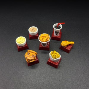 May include: Miniature food items including a cup of soda with a red straw, a bucket of fried chicken, a biscuit, fries, a drumstick, mashed potatoes, and a cinnamon roll. Each item is on a small red square.