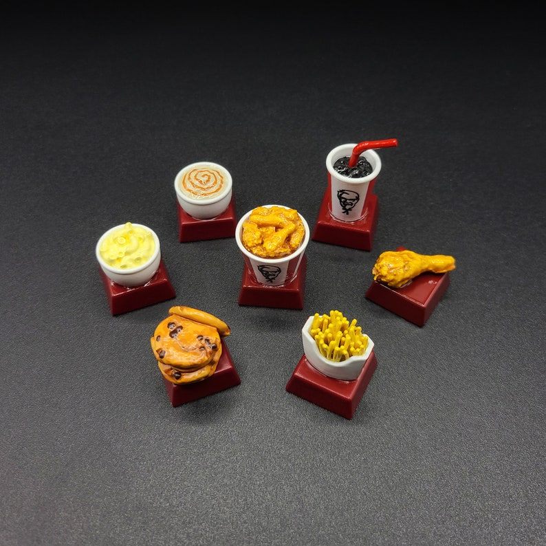 May include: Miniature food items including a cup of soda with a red straw, a bucket of fried chicken, a biscuit, fries, a drumstick, mashed potatoes, and a cinnamon roll. Each item is on a small red square.