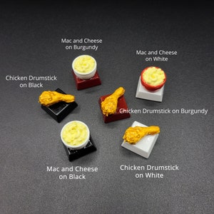 May include: Miniature food items including mac and cheese and chicken drumsticks. The mac and cheese is in small white or red bowls. The chicken drumsticks are golden brown. The items are on black, burgundy, and white platforms. Text on the image reads "Mac and Cheese" and "Chicken Drumstick".