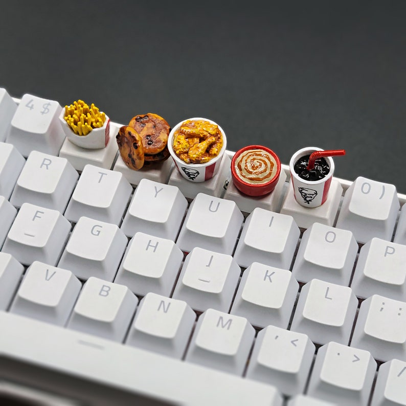 May include: A white mechanical keyboard with custom keycaps shaped like fast food items. The keycaps include a container of fries, cookies, a bucket of fried chicken, a cinnamon roll, and a soda with a straw.