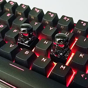 May include: Two black keycaps with a detailed design resembling a helmet. The keycaps are placed on a black keyboard with red backlighting.