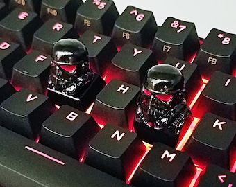 Puede incluir: Dos teclas negras con un diseño detallado que se asemeja a un casco. Las teclas están colocadas en un teclado negro con retroiluminación roja.