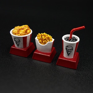 May include: A set of three miniature food items: fried chicken in a bucket, french fries in a carton, and a soda with a straw. Each item features a red and white color scheme and a logo. The items are on red square bases.