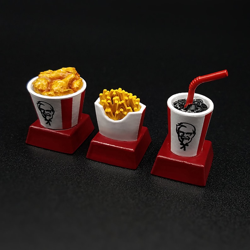 May include: A set of three miniature food items: fried chicken in a bucket, french fries in a carton, and a soda with a straw. Each item features a red and white color scheme and a logo. The items are on red square bases.
