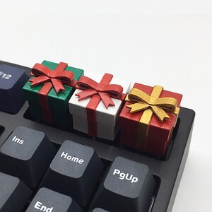 Op de afbeelding: Drie miniatuur geschenkdozen met rode, witte en gouden strikken, die op een zwart toetsenbord liggen met toetsen met de labels "Ins", "Home", "PgUp", "End" en "F12".