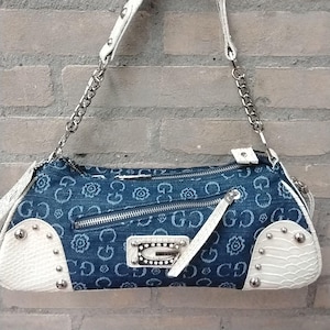 May include: A denim handbag with a white leather handle and accents. The bag features a repeating pattern of the letter "G" and floral designs. Silver chain straps and studded details add to the bag's style. The bag has a zip closure and a white leather accent.