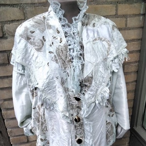 Op de afbeelding: Een wit satijnen jack met een kanten rand en gouden knopen. Het jack heeft een bloemenpatroon en is versierd met franjes.