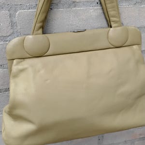 Può includere: Borsa a mano beige chiaro in stile vintage, di forma rettangolare con bordi arrotondati. Ha due manici corti e una chiusura a fermaglio. La borsa, in materiale morbido, è sospesa davanti a un muro di mattoni.