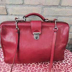 Puede incluir: Un maletín de cuero rojo con asa superior y bandolera. El bolso tiene un cierre de hebilla plateada y herrajes plateados. El maletín está sobre una superficie con estampado de leopardo.