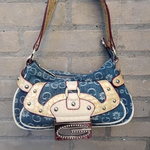 Op de afbeelding: Denim handtas met een bloemenpatroon en de letter "G" op de voorkant. De tas heeft een crèmekleurige flap met zilveren studs en een bijpassende riem. De riem is bruin met bordeauxrode rand.