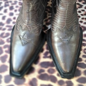 May include: A pair of brown leather cowboy boots with pointed toes and intricate stitching. The boots have a black rubber sole and a pull-on design.