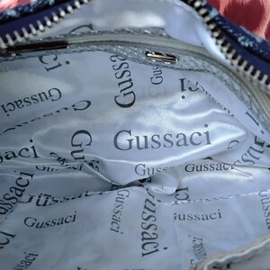 May include: Inside view of a handbag with a silver lining. The lining is printed with the word "Gussaci" in a repeating pattern. The bag has a zip and a blue and leopard print exterior.