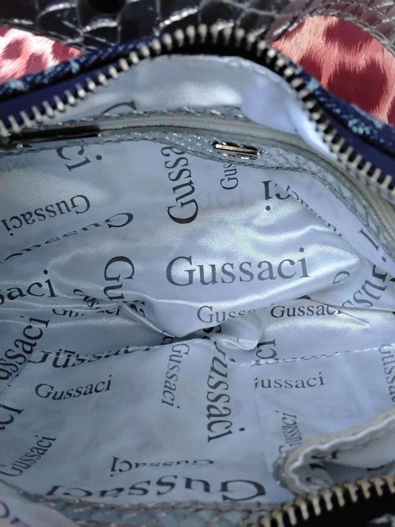 May include: Inside view of a handbag with a silver lining. The lining is printed with the word "Gussaci" in a repeating pattern. The bag has a zip and a blue and leopard print exterior.