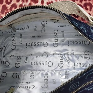 May include: Open denim handbag with a white snakeskin-patterned strap. The interior lining is white with the repeated text "Gussaci". The bag has a black zip and a patterned exterior.