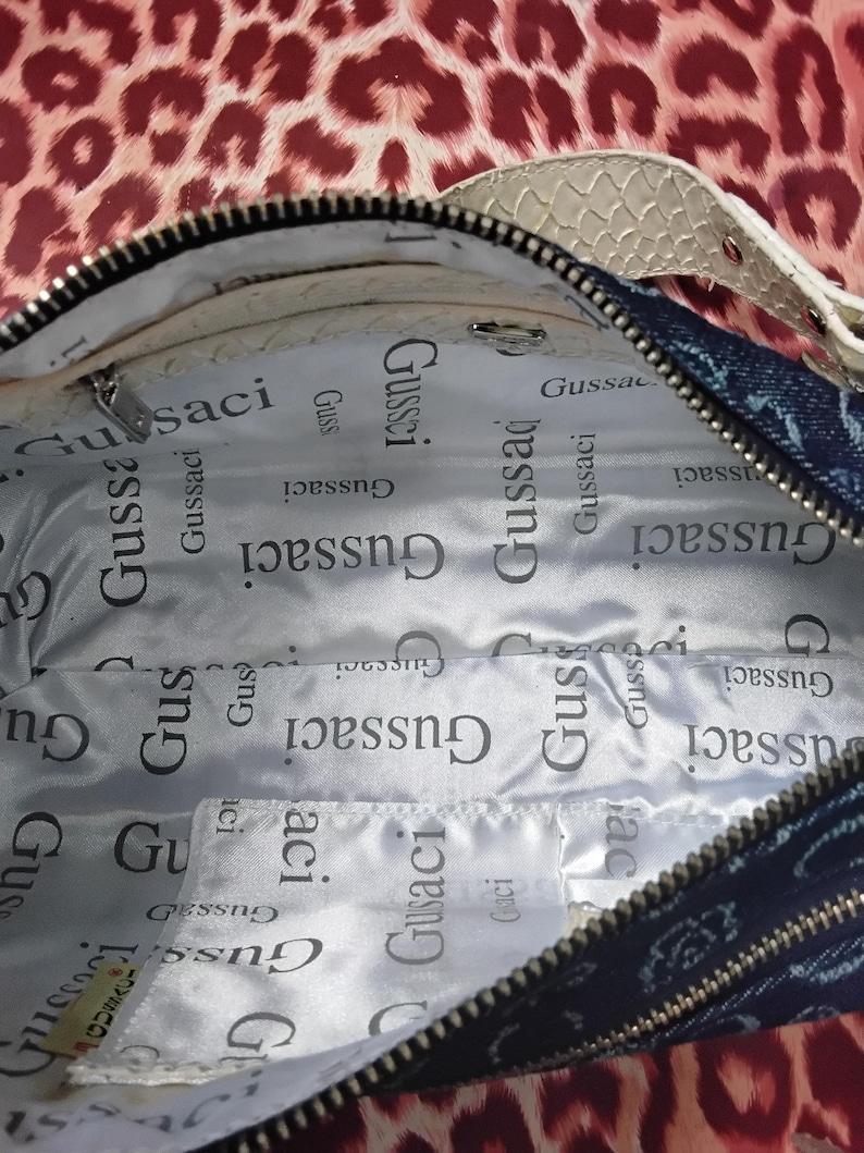 May include: Open denim handbag with a white snakeskin-patterned strap. The interior lining is white with the repeated text "Gussaci". The bag has a black zip and a patterned exterior.