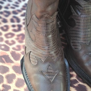 May include: Brown leather cowboy boots with intricate stitching and a pointed toe. The boots have a decorative pattern on the shaft and a black rubber sole.