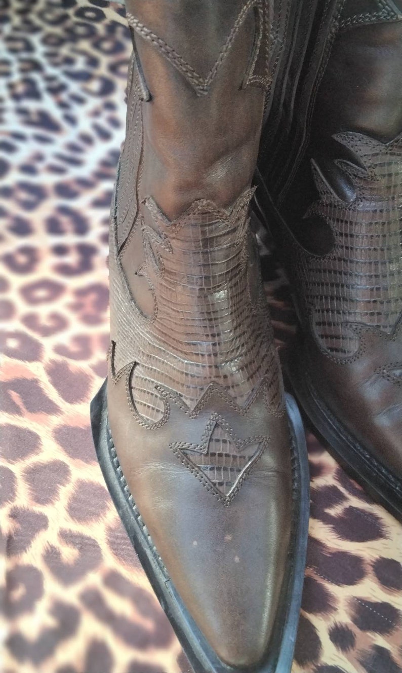 May include: Brown leather cowboy boots with intricate stitching and a pointed toe. The boots have a decorative pattern on the shaft and a black rubber sole.