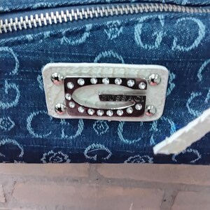 May include: A denim-style handbag with a silver zip and a white leather patch. The patch displays the brand name "GUESS" in silver lettering, encircled by rhinestones and silver studs. The bag has a white strap.