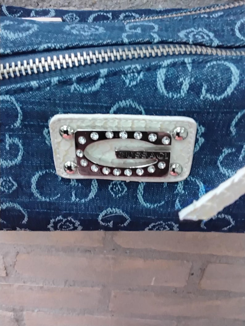 May include: A denim-style handbag with a silver zip and a white leather patch. The patch displays the brand name "GUESS" in silver lettering, encircled by rhinestones and silver studs. The bag has a white strap.
