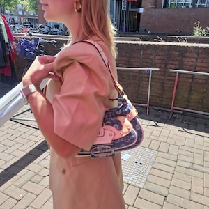 May include: A patterned handbag in blue and pink, with a silver buckle and a white strap. The bag is carried by a person wearing a beige blazer and gold hoop earrings. The background shows a brick wall and a street scene.