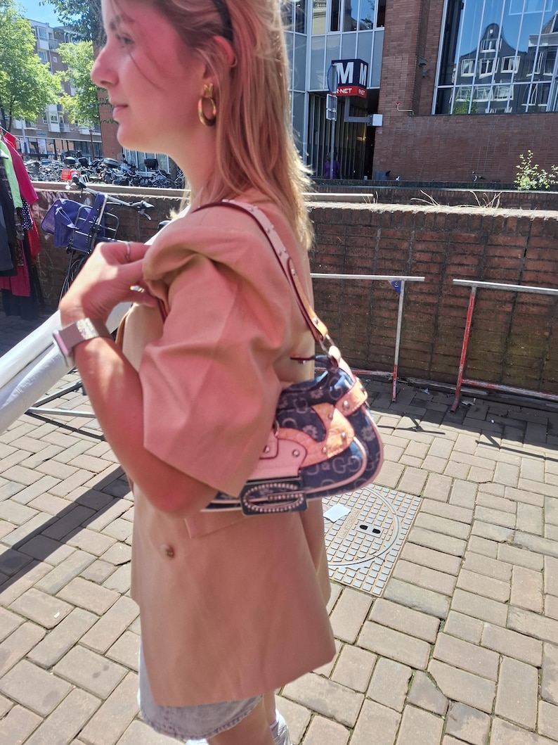 May include: A patterned handbag in blue and pink, with a silver buckle and a white strap. The bag is carried by a person wearing a beige blazer and gold hoop earrings. The background shows a brick wall and a street scene.