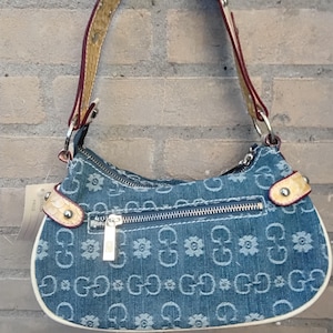 May include: A denim handbag featuring a floral and "G" pattern. The bag has a zip closure, white trim, and a brown and cream strap. It hangs against a brick wall. The bag's strap is 100 cm long.