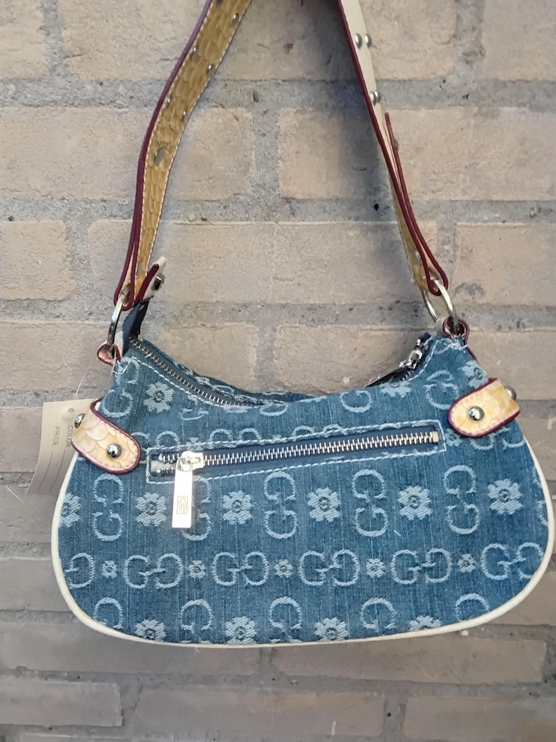 May include: A denim handbag featuring a floral and "G" pattern. The bag has a zip closure, white trim, and a brown and cream strap. It hangs against a brick wall. The bag's strap is 100 cm long.
