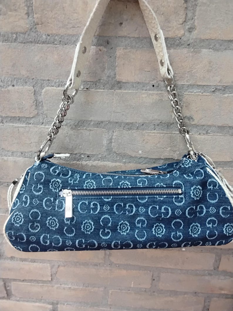 May include: A denim shoulder bag featuring a floral pattern and the letters "CD" repeated. It has a silver chain strap and a white leather handle. A silver zip is across the top. The bag is approximately 30 cm long.