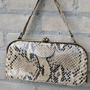 Op de afbeelding: Een rechthoekige handtas met een slangenprint in beige, bruin en zwart. De tas heeft een goudkleurig frame en sluiting, met een bijpassende handgreep. De tas is ongeveer 25 cm breed en 15 cm hoog.