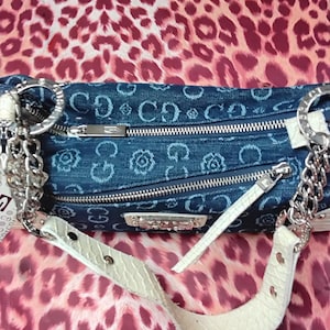 May include: A denim handbag with a repeating floral and logo pattern. The bag has silver-tone chain straps and a white, textured shoulder strap. The background is a pink and red leopard print.