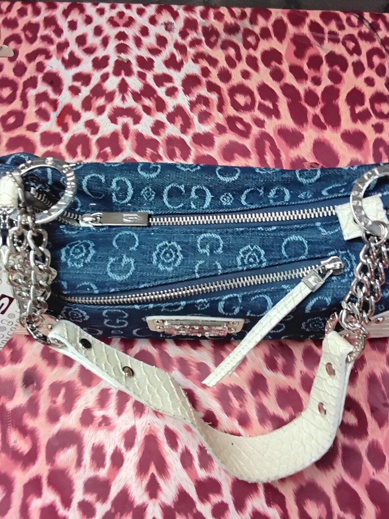 May include: A denim handbag with a repeating floral and logo pattern. The bag has silver-tone chain straps and a white, textured shoulder strap. The background is a pink and red leopard print.