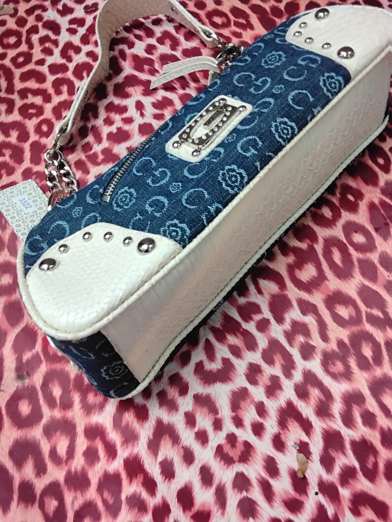 May include: A denim and white handbag with a silver chain strap. The bag has a blue denim top with a floral pattern and a white textured base. Silver studs and a silver logo plate add detail.