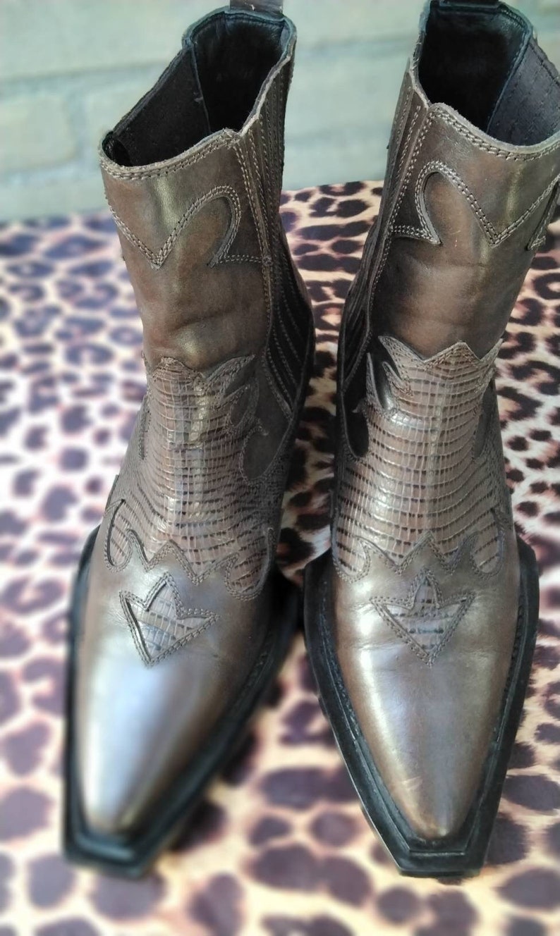 May include: A pair of brown leather cowboy boots with pointed toes and intricate stitching. The boots have a black rubber sole and a pull-on design.