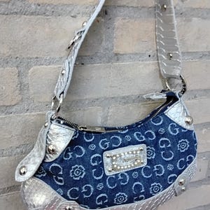 May include: A denim handbag with a silver strap and trim. The bag features a repeating pattern of the letter "G" and floral designs. A silver plaque with rhinestones is centered on the front.