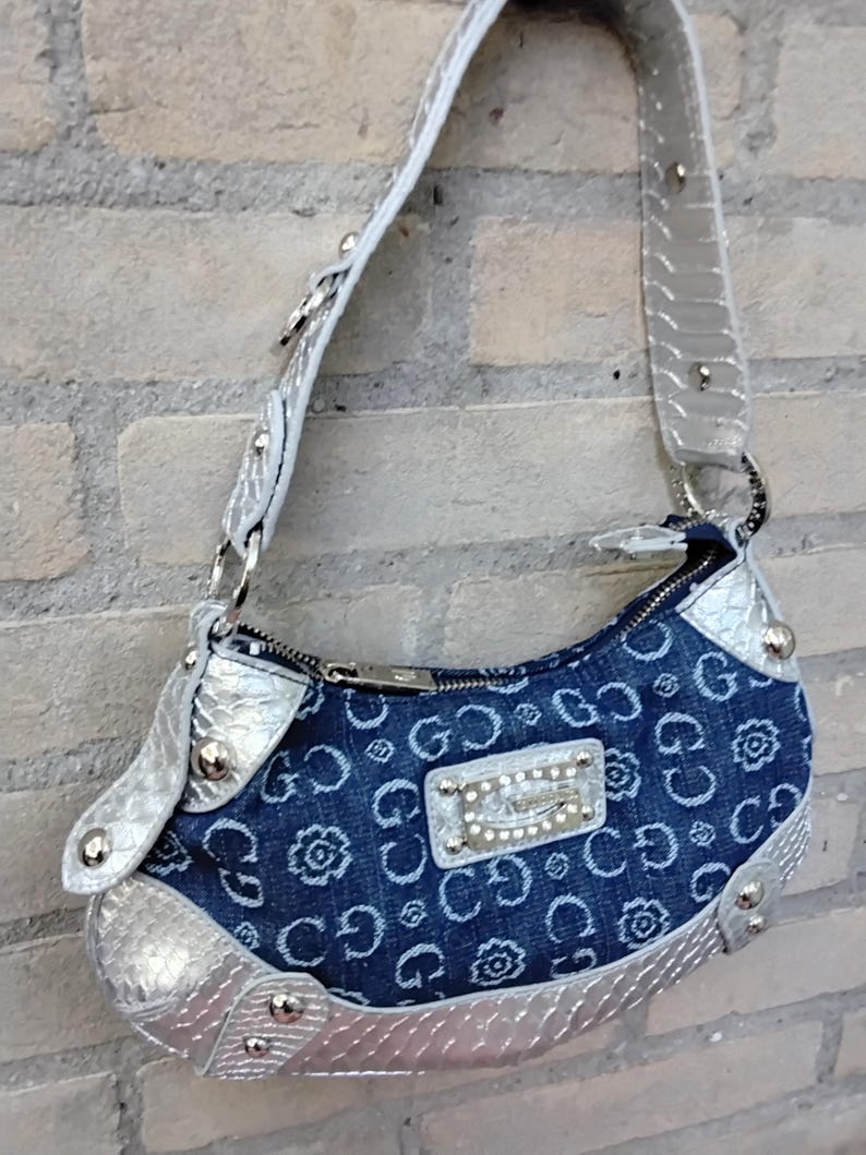 May include: A denim handbag with a silver strap and trim. The bag features a repeating pattern of the letter "G" and floral designs. A silver plaque with rhinestones is centered on the front.