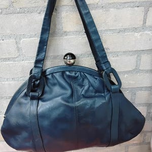 May include: A dark blue leather handbag with a rounded shape and a silver-coloured clasp. The bag has two handles and a structured design. The bag is approximately 30 cm wide and 20 cm tall.