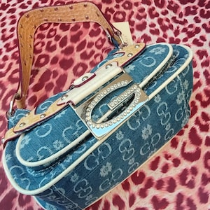 May include: A denim handbag with a repeating pattern of the letters "G" and floral designs. The bag features a silver-tone buckle adorned with rhinestones, a tan leather strap with metal studs, and white trim. The bag is set against a leopard print background.