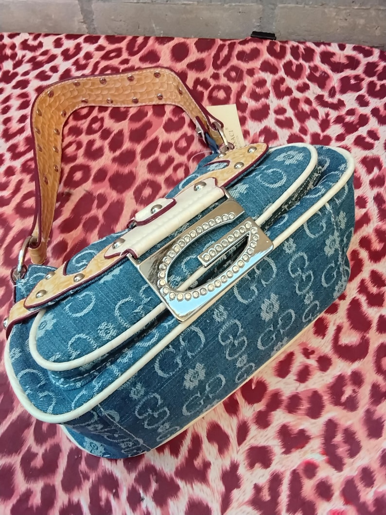 May include: A denim handbag with a repeating pattern of the letters "G" and floral designs. The bag features a silver-tone buckle adorned with rhinestones, a tan leather strap with metal studs, and white trim. The bag is set against a leopard print background.