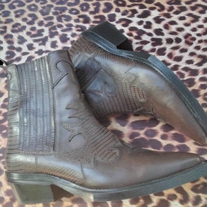 May include: A pair of brown leather cowboy boots with a pointed toe and a low heel. The boots have a decorative stitching pattern and a textured leather design.