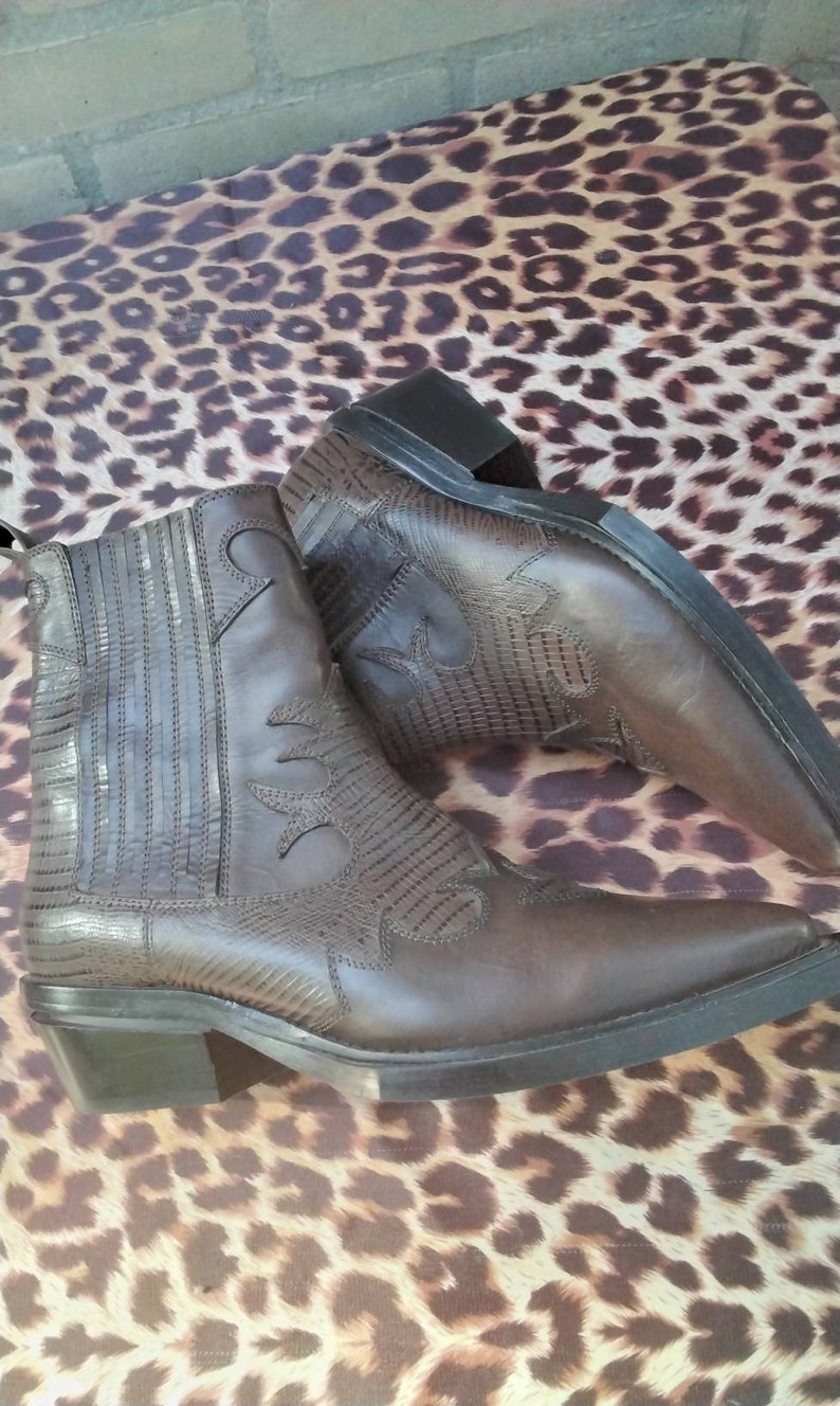 May include: A pair of brown leather cowboy boots with a pointed toe and a low heel. The boots have a decorative stitching pattern and a textured leather design.
